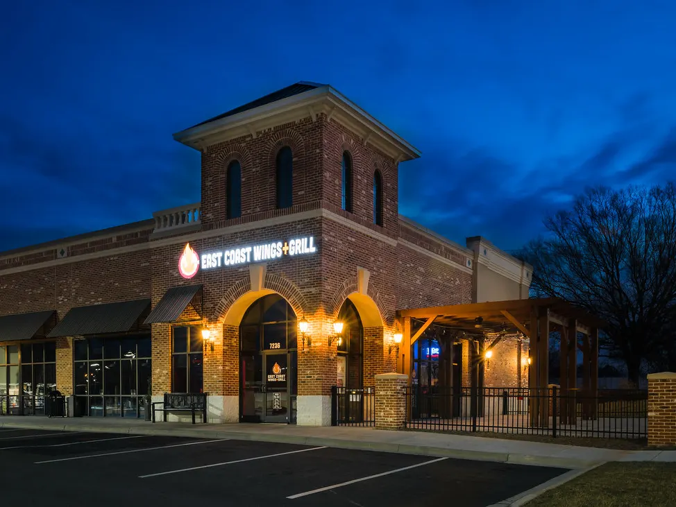 Lit storefronts and a restaurant patio in a Harrisburg shopping center at dusk